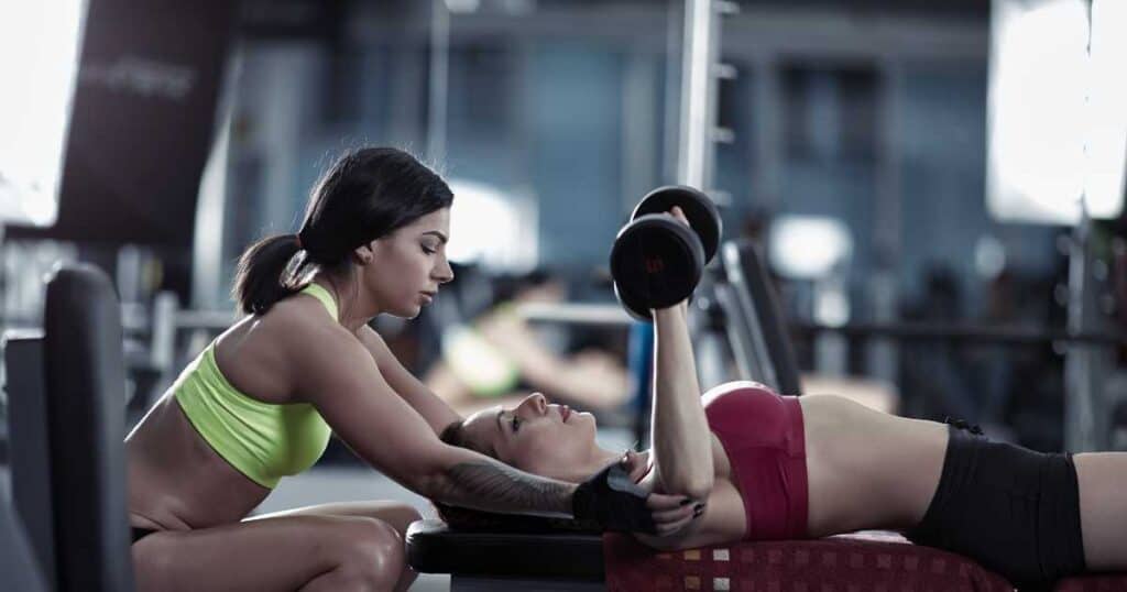 female personal trainer in green sports bra assisting woman lifting dumbbells