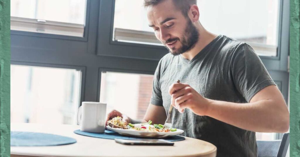man sitting at a table near large window with coffee mug eating a meal with a fork.