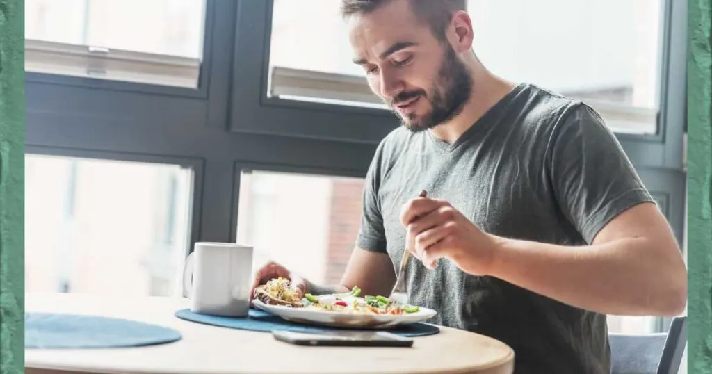 man sitting at a table near large window with coffee mug eating a meal with a fork.