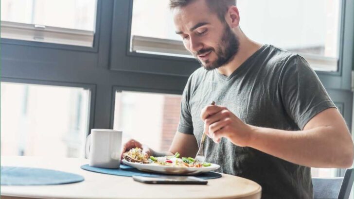 man sitting at a table near large window with coffee mug eating a meal with a fork.