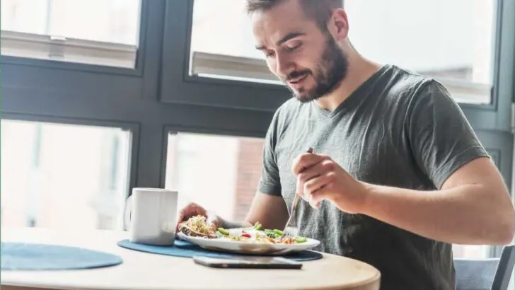 man sitting at a table near large window with coffee mug eating a meal with a fork.