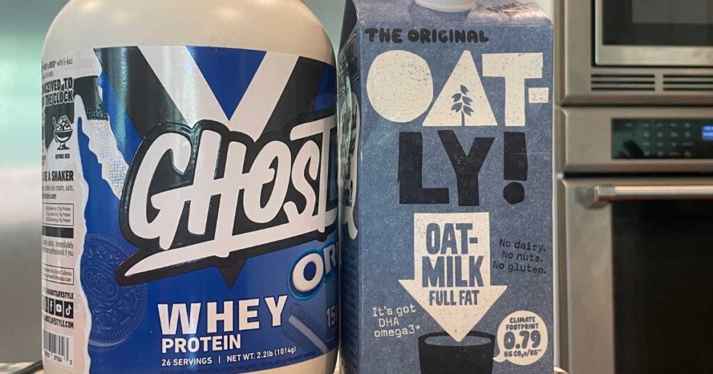 Large container of Ghost oreo whey protein next to a container of Oatly oat milk microwave in background