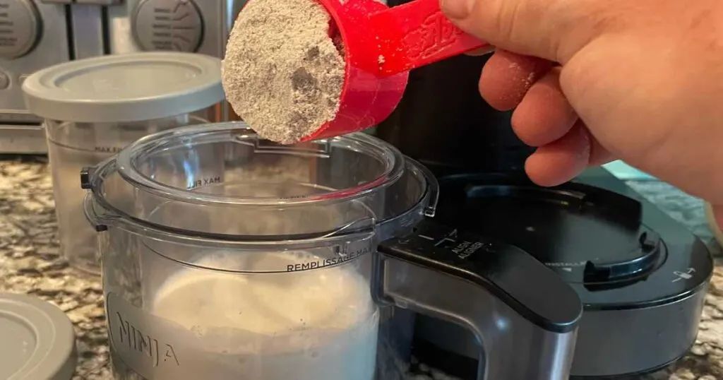hand using a red scoop of protein powder being poured into a ninja container of milk