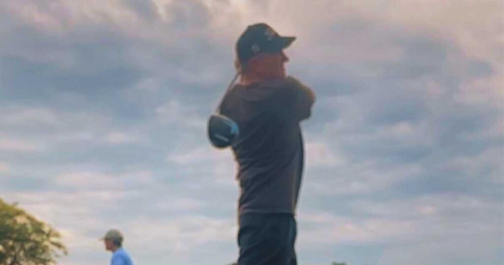 Man wearing a baseball hat swinging a golf club with clouds in background.