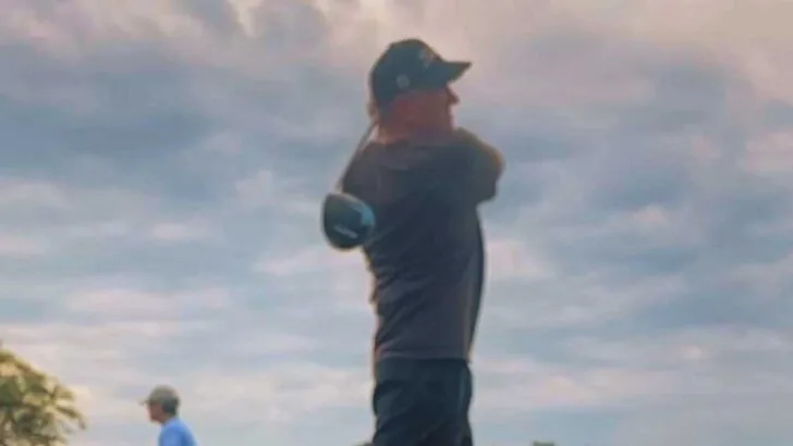 Man wearing a baseball hat swinging a golf club with clouds in background.
