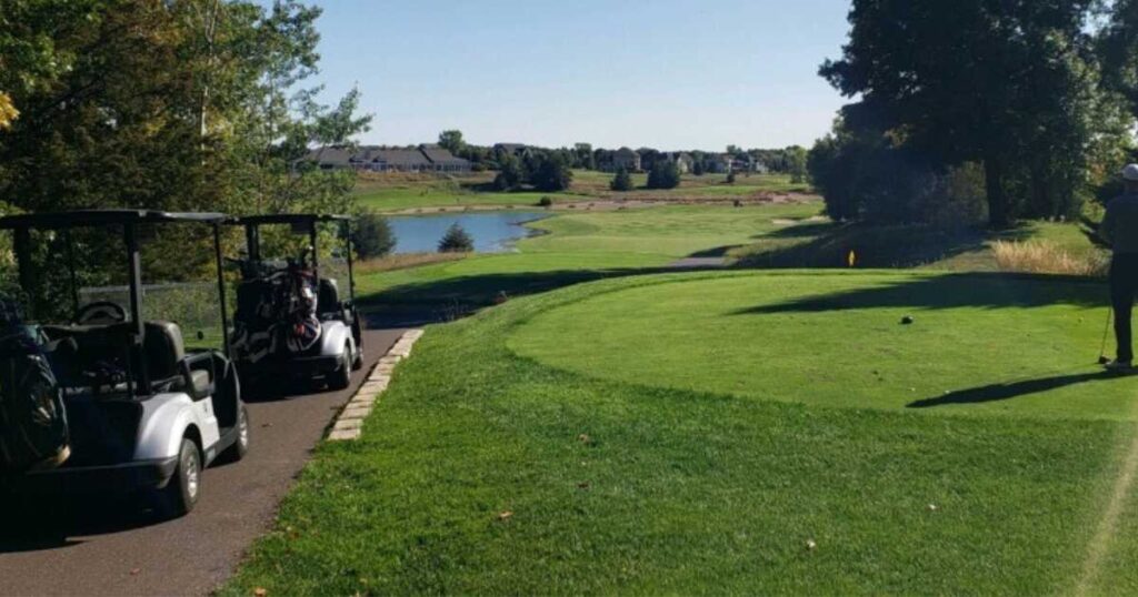 two golf carts on a golf course with a pond and houses in background.
