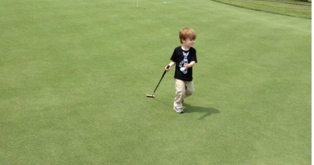 little boy wearing a t-shirt with a tie holding a golf ball and dragging a golf club on a green.