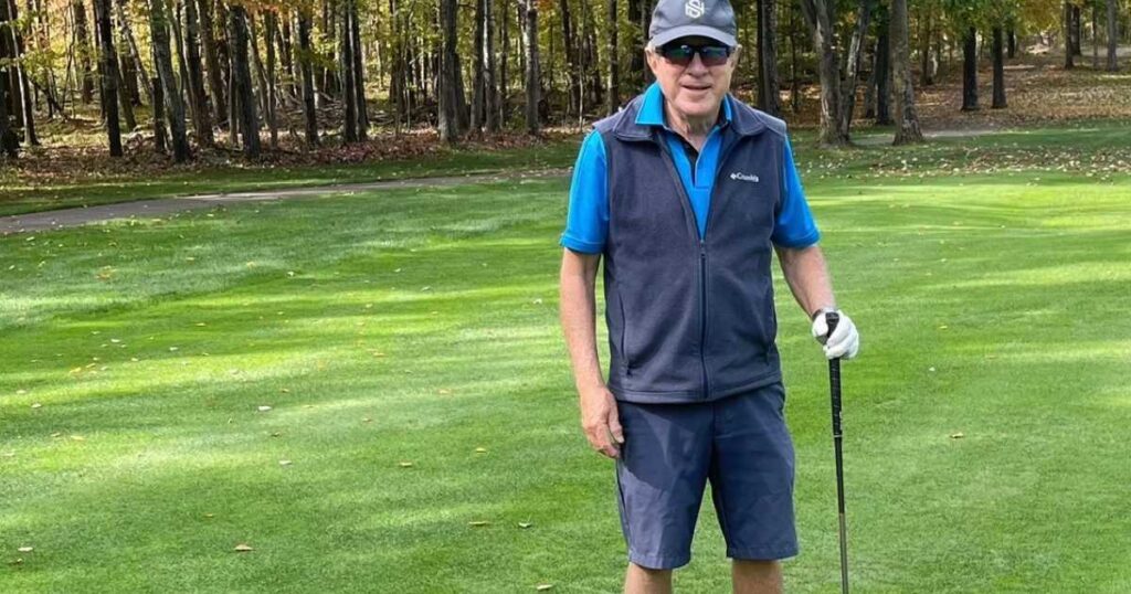older man wearing a baseball cap, vest, golf shirt, and shorts, leaning on a golf club on the greens of a golf course.